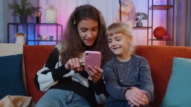 Genç kız kardeşler başarıyı kutluyor. Akıllı telefondan online alışveriş yaparken çığlıklar atıyorlar. Mutlu kardeşler, çocuklar ya da en iyi arkadaşlar boş zamanlarını oyun odasında sosyal medya uygulamalarında geçirirler.
