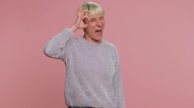 Kısa yeşil saçlı neşeli bir kadın onaylı geri bildirim jesti göstererek kameraya bakıyor. Pembe arka planda genç bir kız. İnsanlar samimi duygular