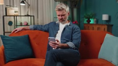 Kanepede oturan sakallı, orta yaşlı bir adam gece evinde cep telefonuyla gülümsüyor. Olgun erkek akıllı telefon sosyal medya uygulamalarında mesajlaşır, rahatlama filmi izler.