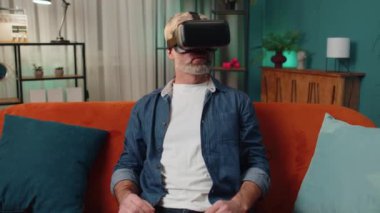 Son sınıf öğrencisi sanal gerçeklik teknolojisi VR uygulama kulaklığı kullanarak 3D 360 video oyunu simülasyonu yapıyor ve modern ev dairesinde film izliyor. Kanepede oturan gözlüklü adam.