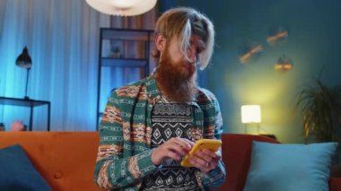 Kanepede oturan hippi kızıl sakallı adam gece evde cep telefonuyla gülümsüyor. Akıllı telefon sosyal medya uygulamaları üzerinden rahatlama filmi izlerken mesajları paylaşan genç adam