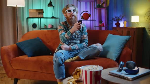 Ενθουσιασμένος Γενειοφόρος Hippie Νεαρός Άνδρας Κάθεται Στον Καναπέ Χρήση Τηλεχειριστήριο — Αρχείο Βίντεο