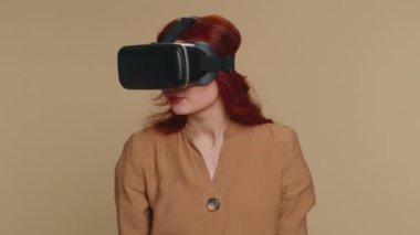 Kızıl saçlı kadın çevrimiçi oyun uygulaması oynamak için kulaklık kullanıyor. Sanal gerçeklik 3D 360 video içeriği izliyor. VR gözlüklü bir kız stüdyo bej arka planında izole edilmiş. Gelecekçi teknoloji