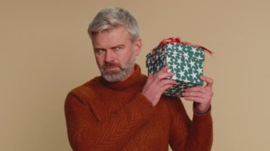 Mutlu orta yaşlı bir adam hediye kutusunu açıyor, doğum günü sürprizini açıyor ve en iyi hediye ikramiyesinden memnun kaldığını belirtiyor. Bej renkli stüdyo arka planında izole edilmiş yaşlı bir adam.