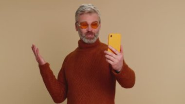 Güneş gözlüklü, orta yaşlı bir gezgin blogger, akıllı telefondan selfie çekiyor, aile veya arkadaşlarla çevrimiçi video görüşmesi yapıyor, canlı çeviri akışı yapıyor. Bej arkaplanlı yaşlı bir adam.