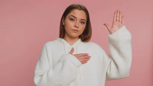 I swear to be honest. Sincere responsible young woman raising hand to take oath, promising to be honest and to tell truth, keeping hand on chest. Caucasian girl isolated on studio pink background