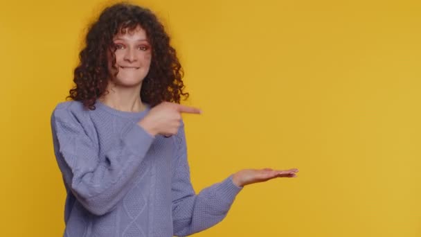 親指を表示し 空の場所を指す素敵な女性 商業テキストの広告エリア 商品プロモーションのためのコピースペース 若い巻き毛の女の子 黄色の壁の背景に撮影室内スタジオ — ストック動画