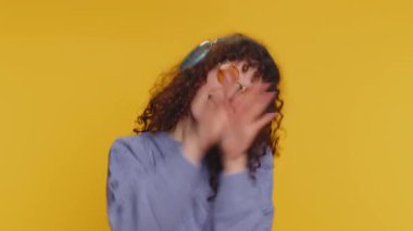 Kıvırcık saçlı, kulaklıklı, müzik dinleyen, dans eden, eğlenen, el kol hareketi yapan, partide rahatlayan mutlu bir kadın. Beyaz kız stüdyo sarı arka planda izole edilmiş.