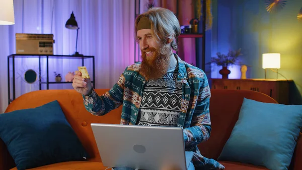 Bearded redhead man using credit bank card and laptop computer while transferring money, purchases online shopping, order food delivery at evening home. Young hippie guy on couch at home evening room