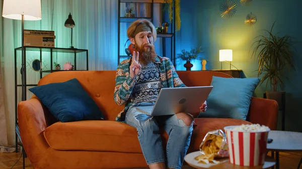Bearded hippie redhead man sitting on couch looking at camera, making video webcam conference call with friends or family, enjoying pleasant conversation. Young guy laughing waving hello at night home