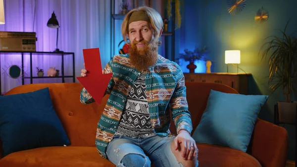 Upset bearded hippie man at home couch showing red arrow pointing down, concept of downgrade, unsuccessful business, fall of stock market money exchange rate bankruptcy fail. Hipster guy in night room