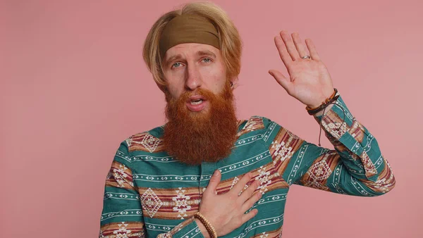 I swear to be honest. Sincere responsible hippie redhead bearded man raising hand to take oath, promising to be honest and to tell truth, keeping hand on chest. Hipster ginger guy on pink background
