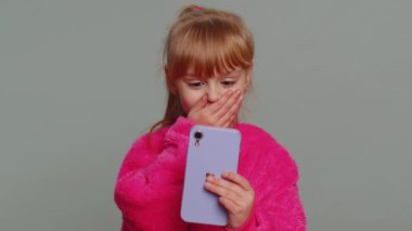 Young preteen child girl kid use mobile smartphone typing browsing say Wow yes found out great big win good news celebrate doing winner gesture. Little toddler children on studio gray wall background