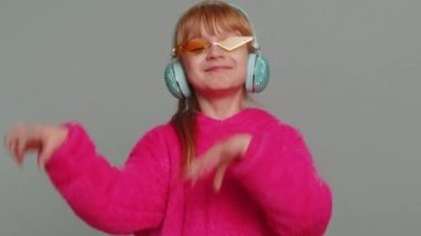 Happy blonde preteen child girl kid listening music via headphones, dancing disco fooling around having fun expressive gesticulating hands. Little toddler children isolated on studio gray background