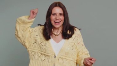 Happy joyful pretty woman in cardigan shouting, raising fists in gesture I did it, celebrating success, winning, birthday, lottery jackpot goal achievemen. Adult girl indoors on gray studio background
