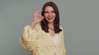 Ok. Joyful happy pretty woman in cardigan looking approvingly at camera showing approve feedback gesture like sign positive something good. Young girl on gray studio background. People sincere emotion
