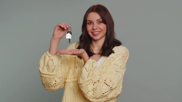 新しい家の家のアパートの鍵を示す手を持ち上げるカーディガン不動産エージェントでかなり若い女性 不動産を借りる 住宅ローン 灰色のスタジオの背景に隔離された千年紀の女の子 — ストック動画
