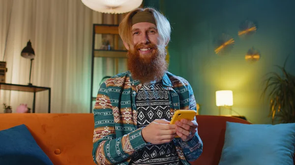 Hipster redhead bearded man sitting on sofa uses mobile phone smiles at night evening home. Young guy texting share messages content on smartphone social media applications online watching relax movie
