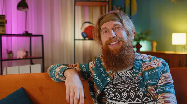 Happy calm bearded hippie redhead man at home couch smiling friendly, glad expression looking away dreaming resting, relaxation feel satisfied concept good news, celebrate win. Young guy in night room