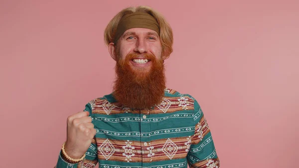 Happy hippie redhead man in pattern shirt shouting, raising fists in gesture I did it, celebrating success, winning, birthday, lottery goal achievemen. Hipster guy boy indoor on pink studio background