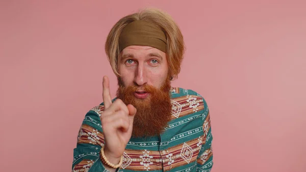Hippie redhead bearded man in pattern shirt shakes finger and saying no, be careful, scolding and giving advice to avoid danger mistake, disapproval sign. Hipster ginger guy on pink studio background
