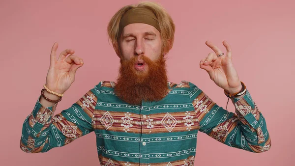 Keep calm down, relax, balance. Hippie redhead bearded man breathes deeply with mudra gesture, eyes closed, meditating with concentrated thoughts, peaceful mind. Hipster ginger guy on pink background