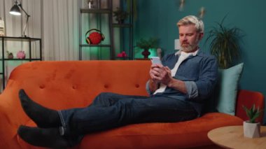 Mature senior bearded man lying on sofa uses mobile phone smiles at night evening home. Mature guy texting share messages content on smartphone social media applications online watching relax movie