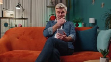 Sad senior middle-aged man use smartphone typing browsing, loses becoming surprised sudden lottery results, bad news, fortune loss, fail, deadline. Elderly mature old guy at evening home room on couch