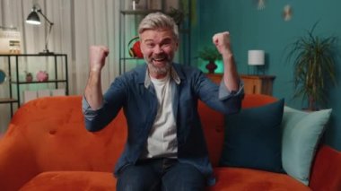 Happy senior man shouting, celebrating success, winning, goal achievement, good news raising fists in gesture I did it, lottery luck at home apartment. Mature middle-aged guy in night room on couch