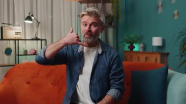 Call me, here is contact number. Middle-aged man looking at camera advertising doing phone gesture like says hey you call me back at home. Mature senior gray-haired old guy at evening room apartment