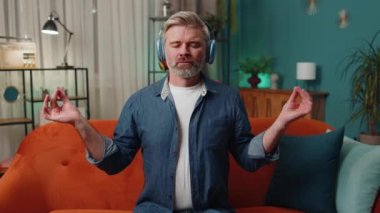 Keep calm down, relax. Middle-aged old man at home couch breathes deeply with mudra gesture, eyes closed meditating with concentrated thoughts, peaceful mind, listen music. Mature guy in evening room
