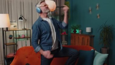Middle-aged mature rocker man in wireless headphones relaxing at home dancing on couch listening energetic disco dancing music, playing on imaginary guitar. People weekend evening leisure activities