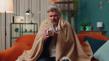 Sick man wrapped in plaid sit alone shivering from cold on sofa drinking hot tea in unheated apartment without heating due debt. Unhealthy senior guy feeling discomfort try to warm up at evening home