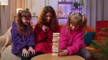 Teenage girl and toddler child sisters build tower from wooden bricks, losing board game competition. Three siblings children kids friends having fun with tower, playing with blocks at home playroom