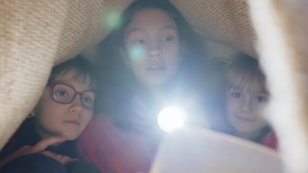 在毛毯格子布罩下的少女和幼儿姐妹们用手电筒阅读有趣的童话书 3个兄弟姐妹 3个孩子 家里的朋友 — 图库视频影像