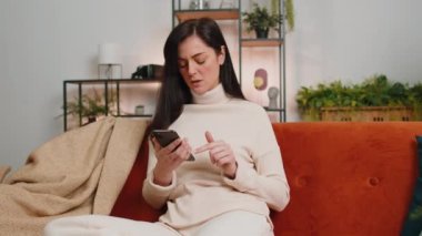 Sad displeased woman use smartphone typing browsing, loses becoming surprised sudden lottery results, bad news, fortune loss, fail, deadline, virus. Young adult brunette girl at home room on couch