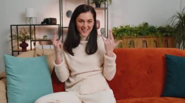 Overjoyed happy young woman showing rock n roll gesture by hands, cool sign, shouting yeah with crazy expression, dancing, emotionally rejoicing in success at home. Adult girl in living room on couch