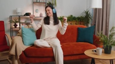 Happy overjoyed brunette woman in wireless headphones relaxing sitting on couch at home apartment choosing listening favorite energetic disco dancing music. People weekend daytime leisure activities