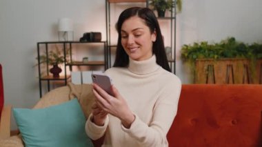 Young woman making smartphone call online video webcam conversation with friends sitting on couch at home in living room apartment. Happy excited girl enjoying mobile loudspeaker talking indoors