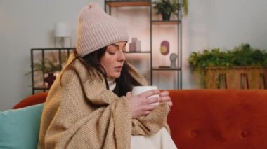 Sick woman wrapped in plaid sits alone shivering from cold on sofa drinking hot tea in unheated apartment without heating due debt. Unhealthy ill girl in hat feeling discomfort try to warm up at home