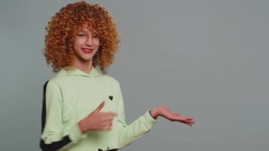 Happy teenager girl showing thumbs up pointing empty place, advertising area for commercial text, copy space for goods promotion banner, copy-space. Young child kid with curly hair on gray background