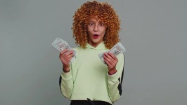 Rich pleased young teenager school girl in glasses waving money dollar cash banknotes bills like a fan, success, lottery winner, big income, wealth. Child kid with curly hair on studio gray background