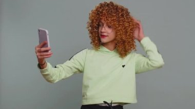 Young child girl kid blogger taking selfie on smartphone holding in hand, communicating video call talking online with subscribers. Teenager children with curly hair isolated on studio gray background