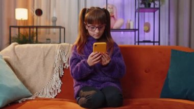 Toddler girl use mobile smartphone typing browsing say Wow yes found out great big win good news celebrate lottery jackpot doing winner gesture. Teen child kid at home in play room sitting on couch