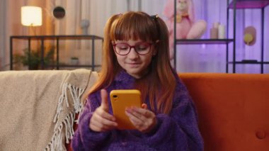 Toddler girl texting share messages content on smartphone social media applications online watching relax movie, news. Teen child, kid at home in night evening living play room sitting on orange sofa