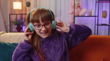 Happy overjoyed toddler girl kid in wireless headphones relaxing sitting on couch at night home choosing listening favorite energetic disco dancing music. People weekend leisure activities. Lifestyles