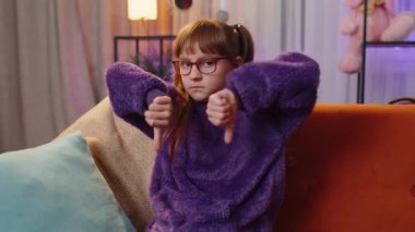 Dislike. Upset girl showing thumbs down sign gesture, expressing discontent, disapproval, dissatisfied bad work at modern home apartment indoors. Displeased teen child, kid in living room on sofa