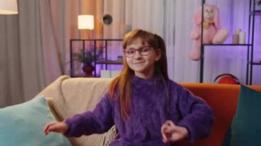Trendy cheerful positive toddler girl having fun dancing, moving to rhythm, dabbing raising hands, making dub dance gesture. Teen child kid at home in night evening living play room sitting on couch