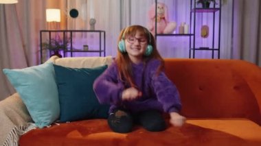 Happy overjoyed preteen toddler child girl in wireless headphones relaxing sits on orange couch at home choosing listening favorite energetic disco dancing music. People weekends leisure activities