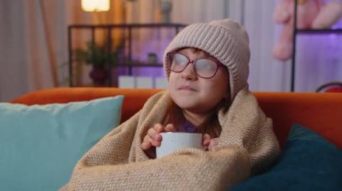 Sick toddler girl wear hat wrapped in plaid sit alone shivering from cold on sofa drinking hot tea in unheated apartment without heating due debt. Unhealthy child feeling discomfort try to warming up